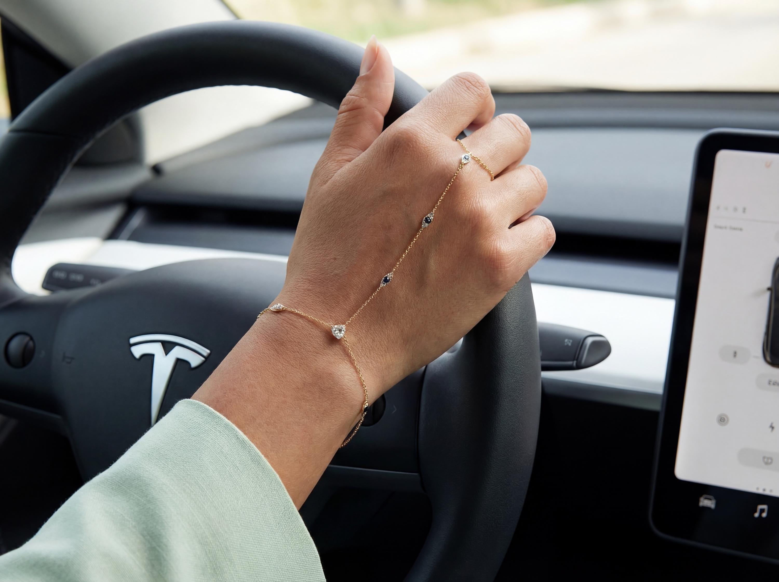 Woman wearing dainty 14k gold evil eye hand chain bracelet while holding car steering wheel, showing minimalist chain connecting finger ring to bracelet with sapphire style stones.