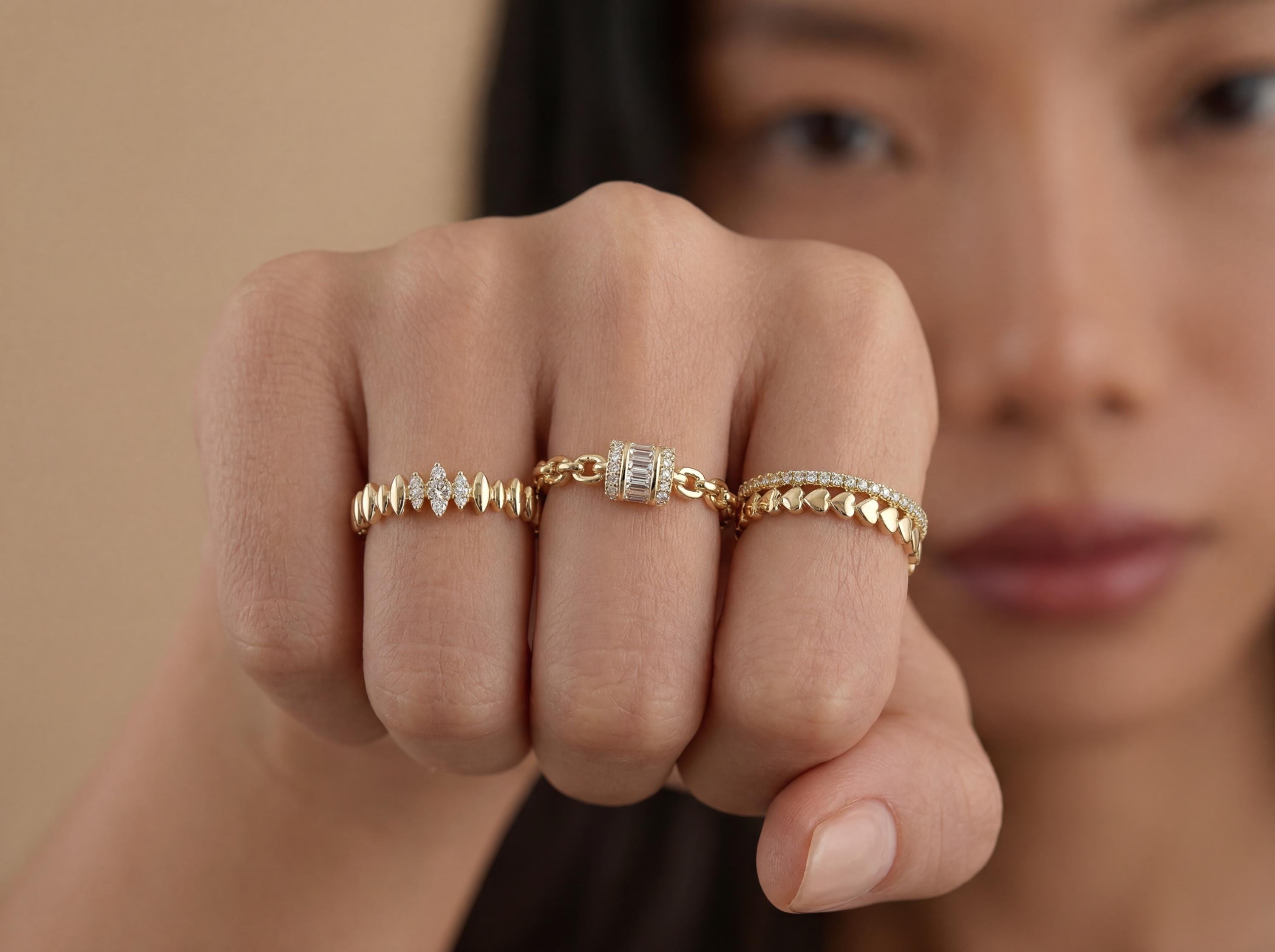 Close view of hand wearing three thin gold stacking rings with heart motif band, chain ring with rectangular diamond detail, and textured diamond wave band.