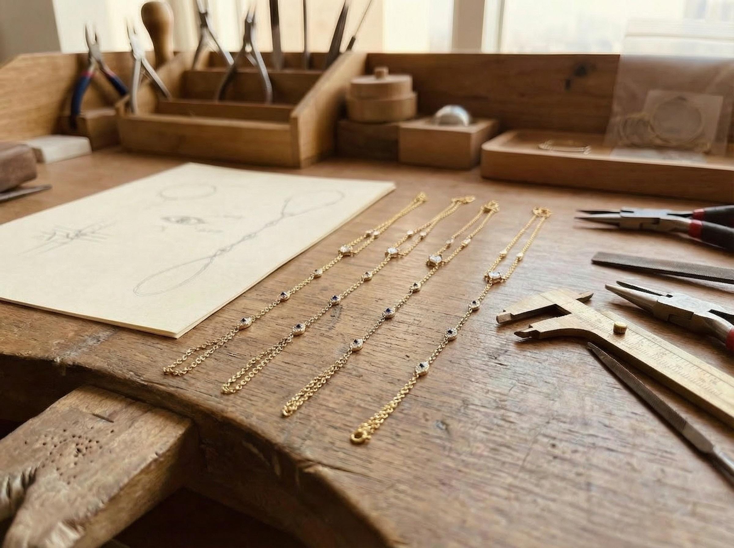 Gold hand chain bracelet with evil eye stones placed on wooden jewelry workbench next to design sketch and professional tools, showing handmade fine jewelry creation process and delicate chain details.