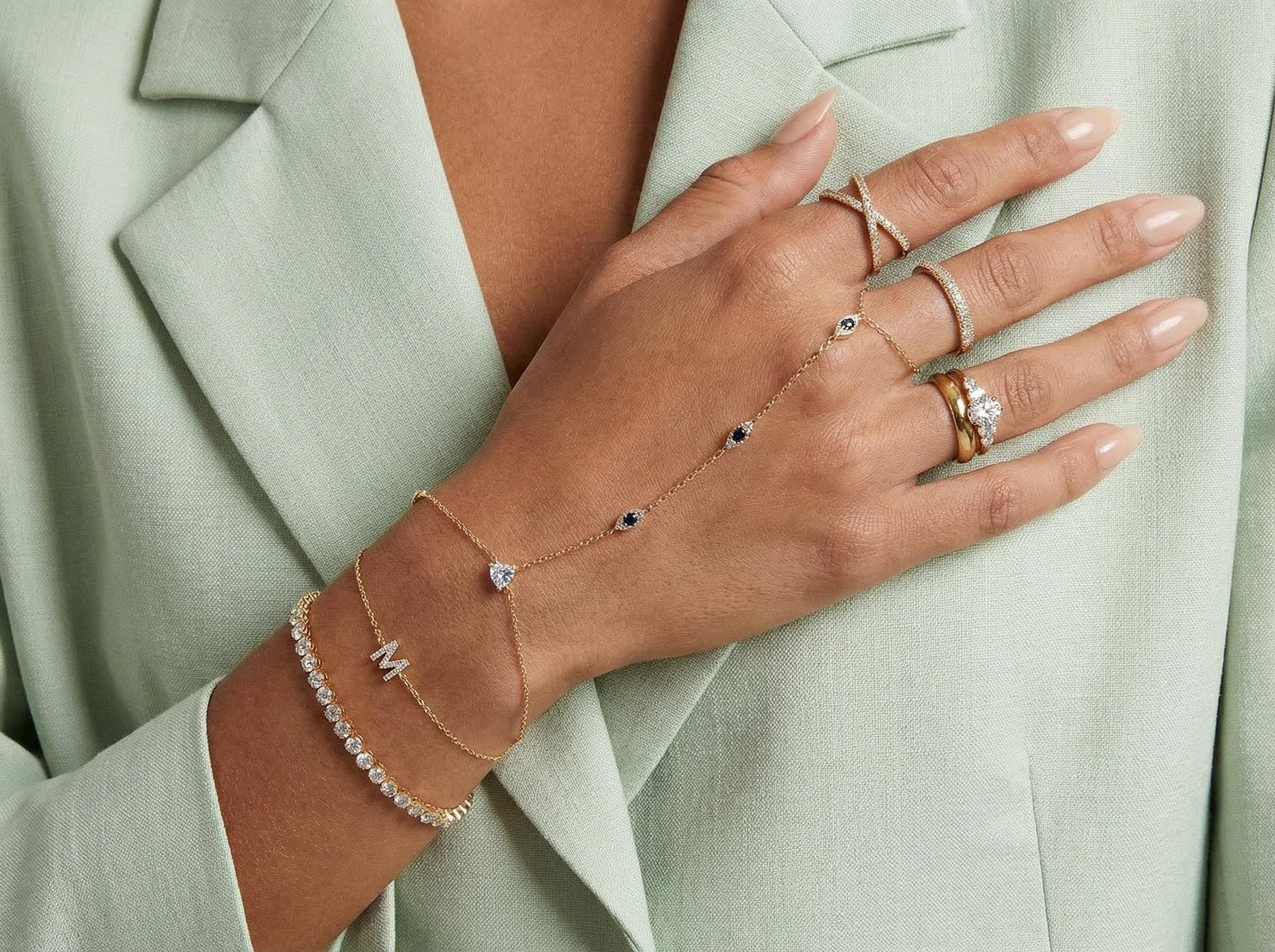 Close-up of woman's hand wearing gold evil eye hand chain bracelet paired with diamond rings and layered bracelets, elegant minimalist fine jewelry styling.