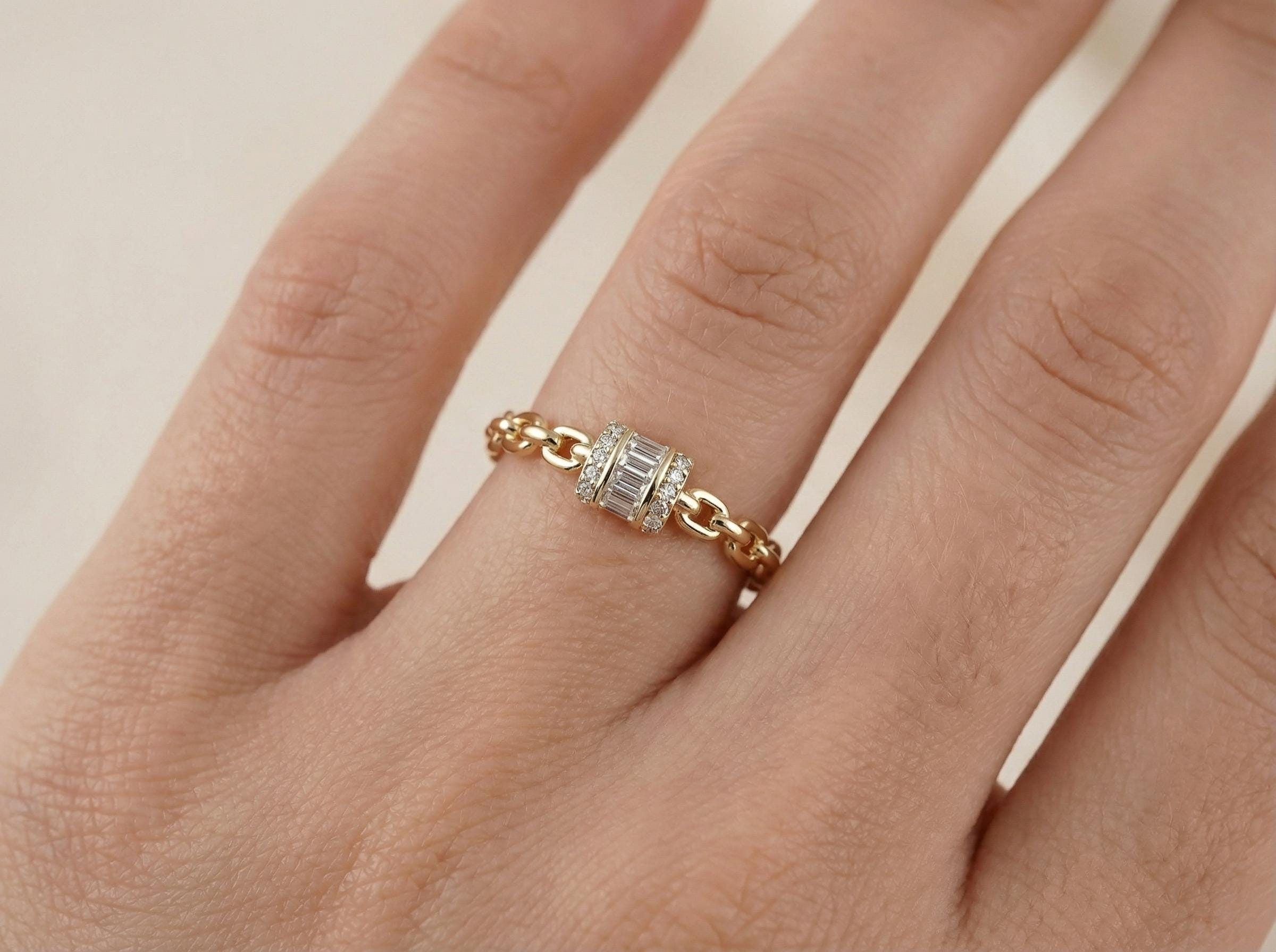 Close-up of gold chain link ring featuring baguette cut diamond center surrounded by small diamonds, worn on finger in minimalist style.