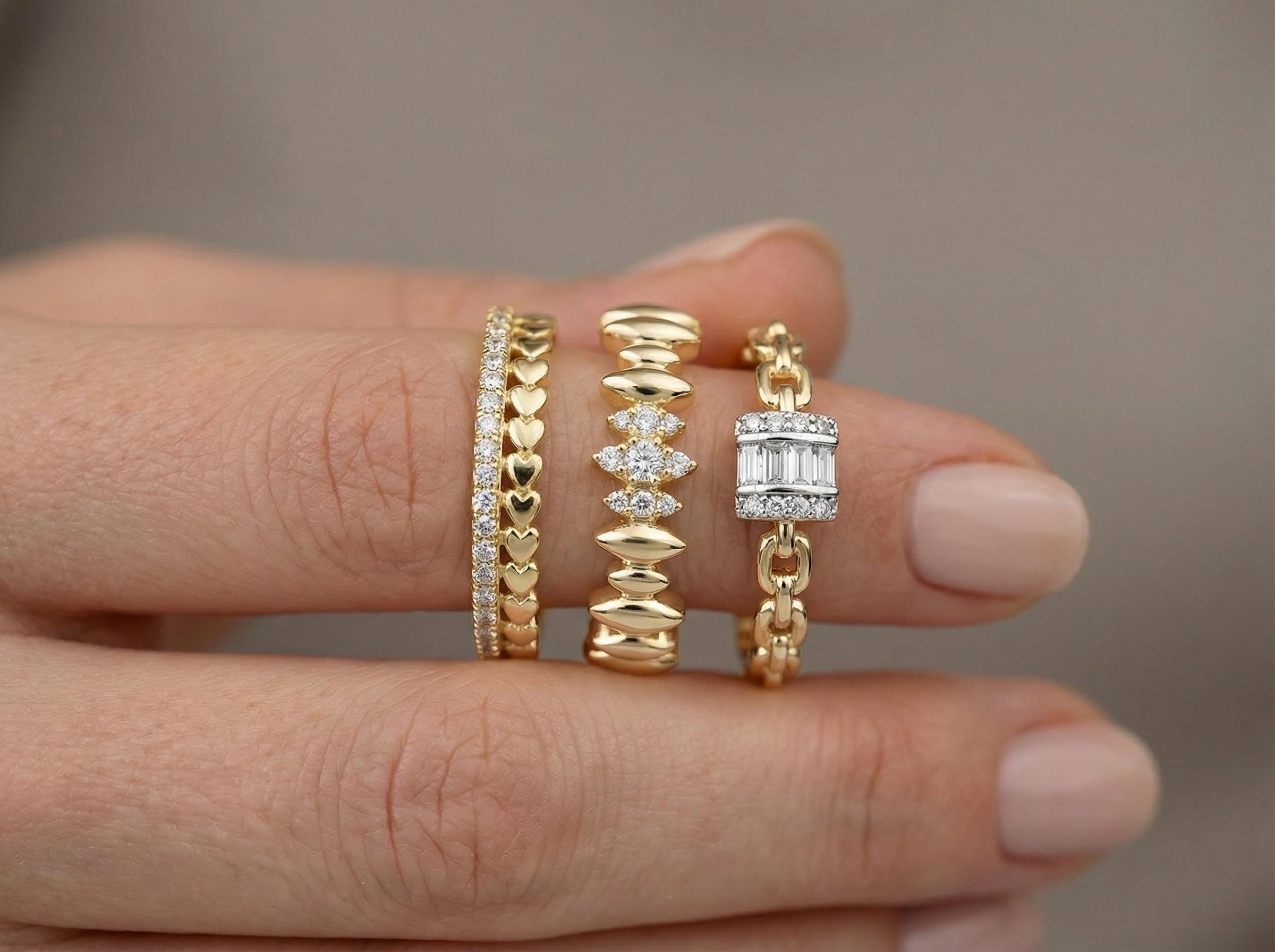 Close-up of three 14k gold rings stacked on finger, featuring heart shape band with small diamonds, baguette diamond chain ring, and sculpted wave band with diamond accents